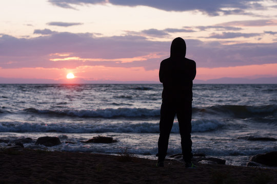 Solitude, loneliness, mindfulness concept. Silhouette of young man standing on edge of beach and looking to horizon of sea waves. Dreaming and thinking alone person at sunset seashore. 
