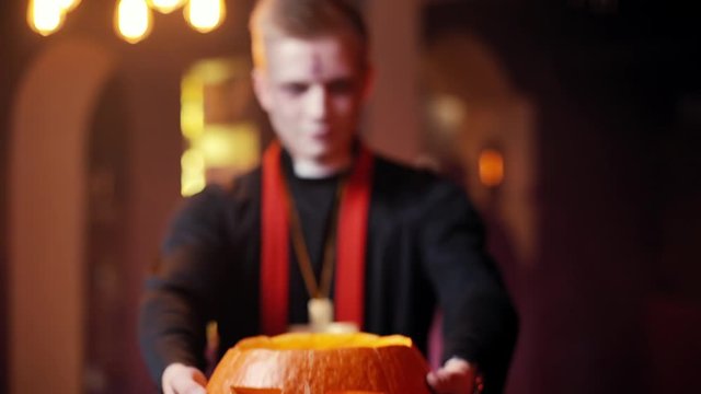 Young man in Halloween priest costume raises carved pumpkin in front of camera. Blurry human background, no visible face. Shot in slow motion