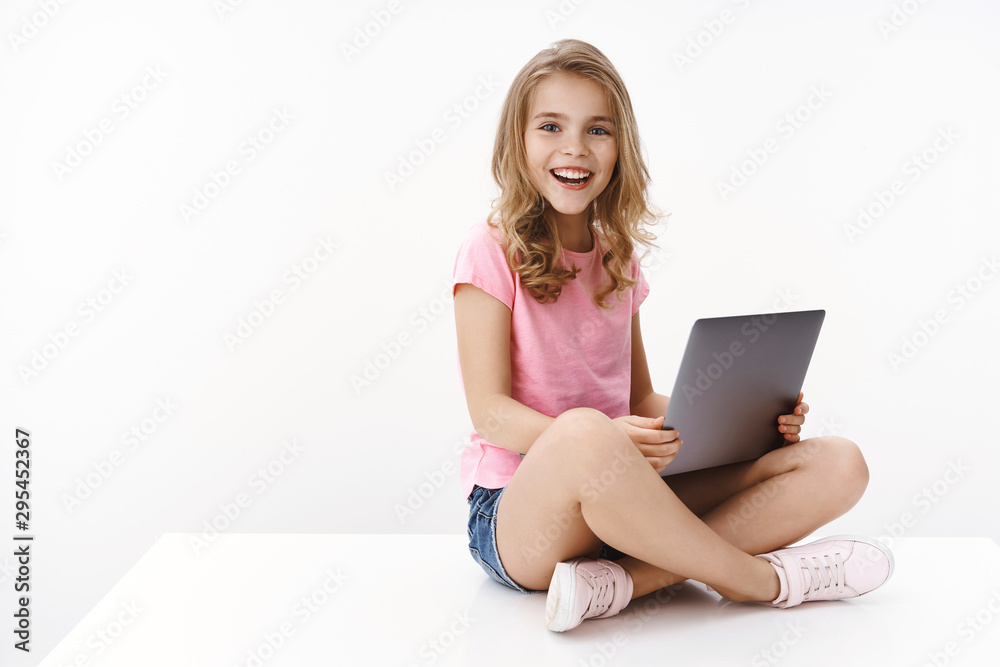 Wall mural pretty cheerful little kid girl sit crossed legs, hold laptop, studying at home, excited learn new i