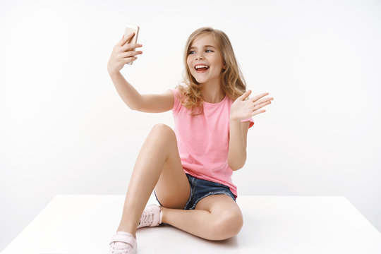 Cheerful enthusiastic happy blond teenage girl sitting crossed legs on floor, hold smartphone, record blog, communicate father abroad, taking selfie, waving hand greeting say hi mobile phone
