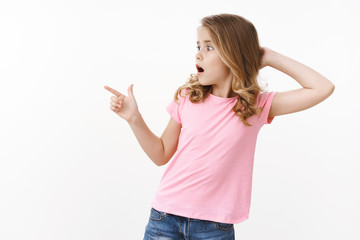 Shocked amazed cute blond little girl kid, touch head stunned, turn pointing left copyspace amazed,...