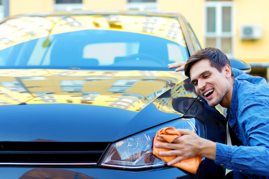 Happy cheerful owner of new black car is wiping, cleaning auto with orange rag. Smiling brunette man driver in denim shirt is hugging and stroking vehicle. In love with automobile concept.