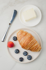 Croissant with berries in white bowl and butter knife on white table. Top view. Healthy breakfast