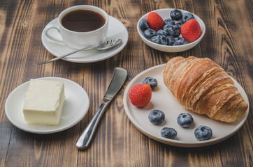 Coffee cup, croissant with berries in white bowl and butter on wooden background. Healthy breakfast with fresh berries