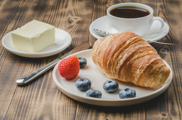 Coffee cup, croissant with berries in white bowl and butter on wooden background. Healthy breakfast with fresh berries