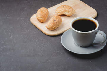 Grey cup with black coffee and wooden tray with croissants on dark table. Copyspace. Coffee break.