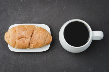 White mug with black coffee and croissant on dark stone table. Top view. Coffee break.