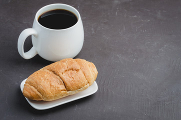 White mug with black coffee and croissant on dark stone background with copyspace.  Coffee break.