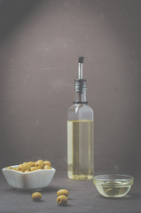 Green olives in a white ceramic bowl and glass bottle of olive oil on a dark stone background. Selective focus. Organic olive oil concept