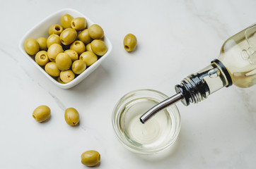Olive oil pouring from bottle in bowl and fresh olives in ceramic plate and on white stone background. Top view.