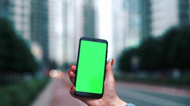 Kyiv - June 5, 2018: Close up hands woman holding use black phone with green screen on busy street modern skycrapers background scrolling pages swiping surfing internet technology smartphone chroma