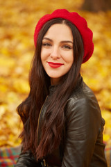 Beautiful woman in autumn park, enjoing warm sunny weather. Close up image of happy dark haired young wearing black leather jacket and red beret, looking with charming smile directly at camera.