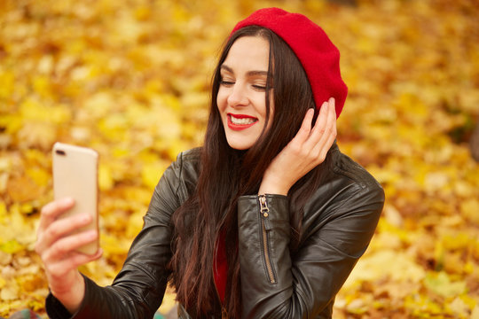 Fashion portrait of young woman outdoor in autumn park or foreest, lady wearing lather jacket, and red beret, taking self picture viaher modern telephone, charming girl surrounded with yellow leaves.