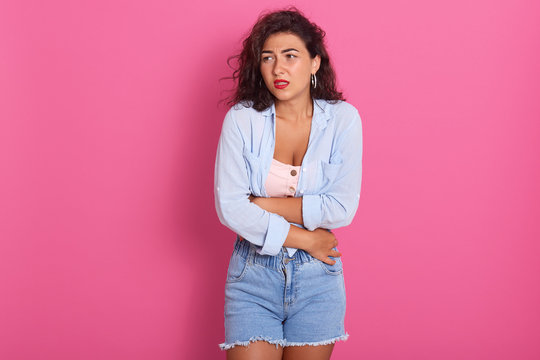 Indoor Shot Of Displeased Caucasian Woman Suffers From Pain In Stomach, Holds Hands On Belly, Looks With Upset Facial Expression, Feels Cold, Wears Casual Clothes, Isolated Over Pink Background.