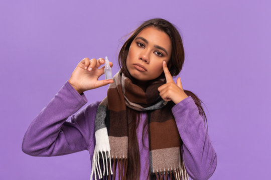Pretty african-american girl in a sweater and cosy checked scarf holding nasal drops to cure common cold standing isolated over purple background. Catching flu in winter.