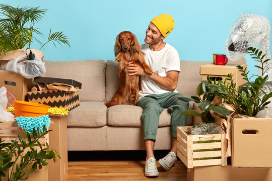 Happy Man Pets Pedigree Dog, Pose On Sofa, Move To New House, Lot Of Packing Cartons Around, Rejoice Buying Modern Flat, Rest After Moving In. Homeowner With Pet Spend First Day At New Dwelling