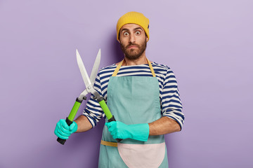 Professional gardener holds trimming shears, busy cutting bushes and pruning trees at home garden, wears apron and gloves, does manintenance work, shows tools for work, takes care of plants.