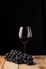 Glass of wine on wooden table. A bunch of ripe black grapes. Black background.
