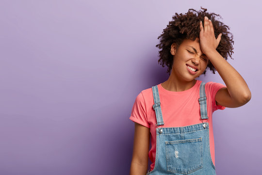 Disappointed troublesome woman keeps palm on forehead, regrets did something wrong, face problematic situation, dressed in fashionable otfit, poses over purple wall, forgets about important task