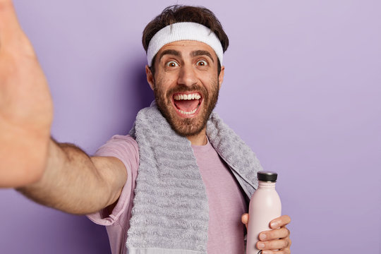 Happy sportsman extends hand with unrecognizable device, takes selfie in gym during workout, holds water bottle, stays hydrated and healthy, being energetic wears towel on neck isolated on purple wall
