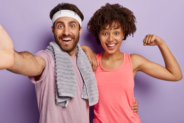 Indoor shot of joyful diverse couple keep muscle flexible, live healthy life, have daily workout, wear sports clothing stand closely look at camera with happy expression show biceps motivated to sport