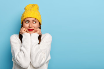 Horizontal shot of happy dreamy European woman wears minimal makeup, red lipstick, looks aside, dressed in yellow hat and white sweater, poses against blue background, being fascinated and pleased