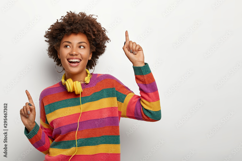 Wall mural Cheerful Afro American woman raises arms and points with index fingers, dances happily to music, wears striped colored sweater and stereo headphones, has overjoyed expression, models indoor. - Wall murals
