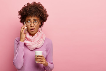 Unhappy stressed shocked Afro American woman talks via mobile phone, holds takeout coffee, hears...