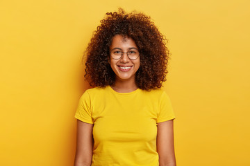 Happy alluring young woman with curly dark hair, looks forward to exciting event, grins joyfully, glad to participate in advertising campaign, wears big round spetacles and yellow t shirt, feels lucky