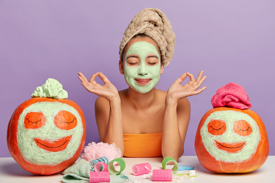 Relaxed woman gets beauty treatments, meditates indoor, poses at table with cosmetic accessories and autumn crops, applies facial masks on pumpkins, has clean healthy skin, isolated over purple wall