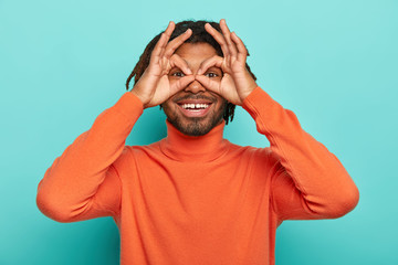 Funny cheerful guy holds hands near eyes, pretends looking through binoculars tries to find love of life, has dreadlocks wears orange turtleneck, shows zero or okay gesture over eyes, isolated on blue