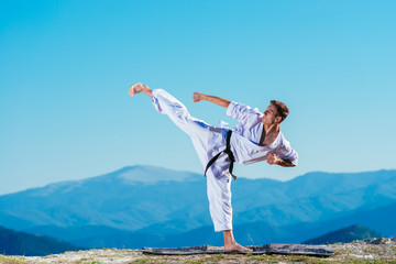 Karate man in a kimono performs a side leg-foot kick(Mae-geri) while standing on the green grass on...
