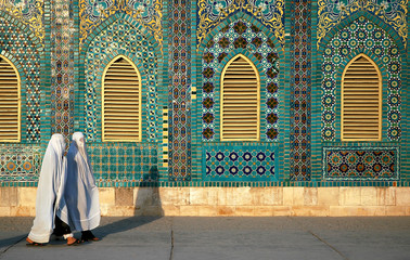 The Blue Mosque in Mazar-i-Sharif, Balkh Province in Afghanistan. Two women wearing white burqas (burkas) walk past a wall of the mosque adorned with colorful tiles and mosaics. Northern Afghanistan. - obrazy, fototapety, plakaty