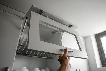 Hand holds the door of the dryer with utensils. Dish drying metal rack with white clean plates. Traditional comfortable kitchen. Open white dish draining closet with wet dishes of plates, bowls.