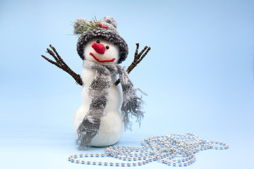 Toy snowman on a blue background. A snowman in a gray hat and a gray scarf stands near a silver beads. Place for text