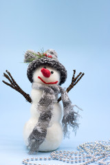 Toy snowman on a blue background. A snowman in a gray hat and a gray scarf stands near a silver beads. Place for text