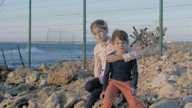two poor suffering orphan refugees children boy and girl sitting on stone near sea with state border with migration razor barbed fence behind