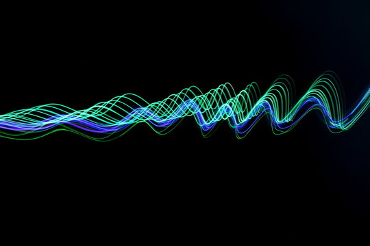 Long exposure photograph of neon green and blue colour in an abstract swirl, parallel lines pattern against a black background. Light painting photography.