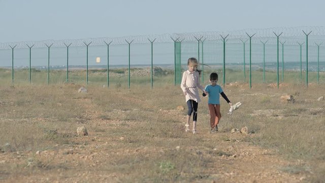 two poor children refugees family girl and boy with toy walking along desert with razor barbed fence on state border