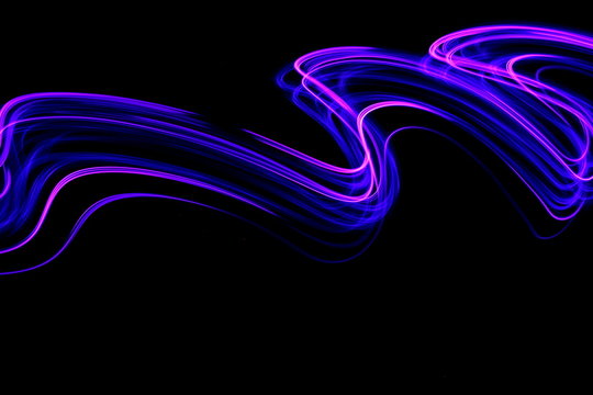 Long exposure photograph of neon pink and purple colour in an abstract swirl, parallel lines pattern against a black background. Light painting photography.