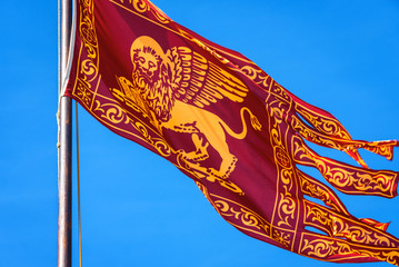 Flag of Venice waving on street in Venice, Italy. Golden winged lion on red flag, closeup view. Old Venetian flag is on background of blue sky in summer.