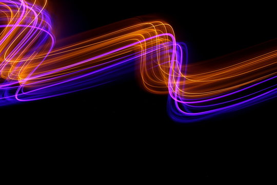 Long exposure photograph of neon purple and gold colour in an abstract swirl, parallel lines pattern against a black background. Light painting photography.