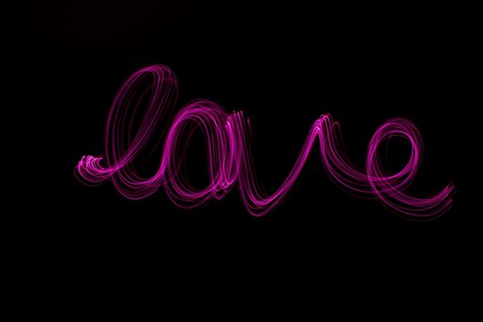 Long exposure photograph of the word 'love'  in pink neon colour in an abstract swirl pattern against a black background. Light painting photography.