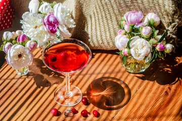 Solemn rustic still life with glass of red drink with autumn berries surrounded by white flowers on textile background on wooden table. Diagonal striped shadow of the blinds on bright sunny day