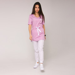 young stylish brunette nurse in pink medical costume with white pants is standing in step, looking straight and smiling cute on the white wall background. medical fashion concept, free space