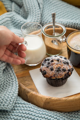 Frau trinkt Kaffee mit Blaubeermuffin, Milch und Zucker auf Holztablett und gemütliches Sofa mit blau Wolldecke und gelb Kissen 