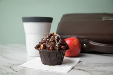 Haselnuss Muffin, Kaffee im to go Becher und Apfel als Frühstück auf dem Weg zur Arbeit mit Leder Aktentasche auf Marmor Tisch und Grün Hintergrund