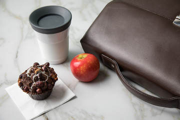 Haselnuss Muffin, Kaffee im to go Becher und Apfel als Frühstück auf dem Weg zur Arbeit mit Leder Aktentasche auf Marmor Tisch und Grün Hintergrund