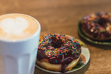 Coffee with a drawn heart and milk on a wooden table in a coffee shop. two chocolate donuts with scattering on the table next to the coffee