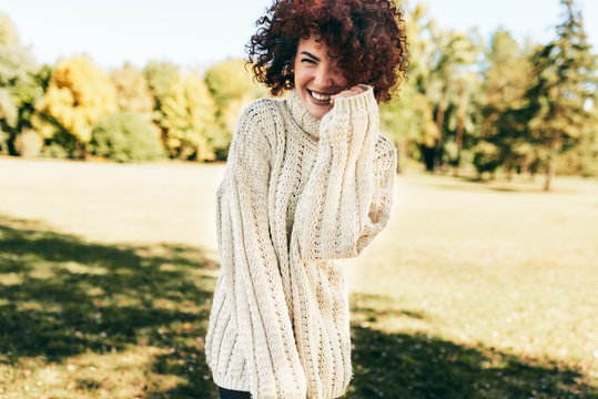 Horizontal image of beautiful young woman smiling and posing against nature background with curly hair, have positive expression, wearing knitted sweater. People, lifestyle